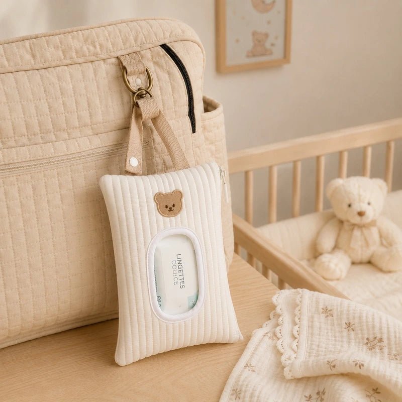 Pochette à lingettes blanche accrochée à un sac à langer près d'un lit bébé, paquet de lingettes visible dans la fenêtre