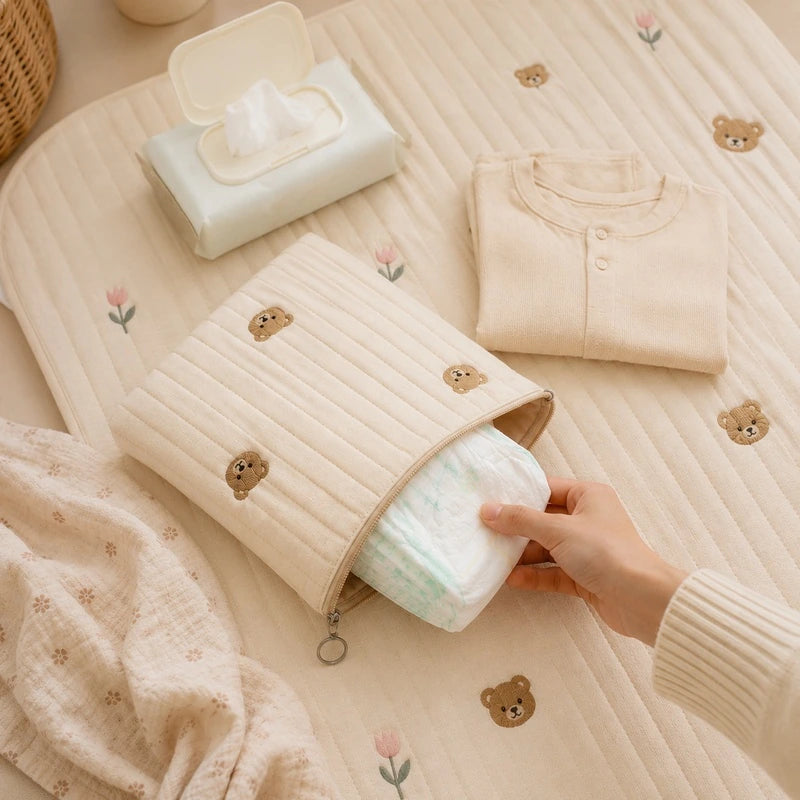 Pochette couche bébé ours ouverte, main y plaçant une couche, entourée de vêtements bébé